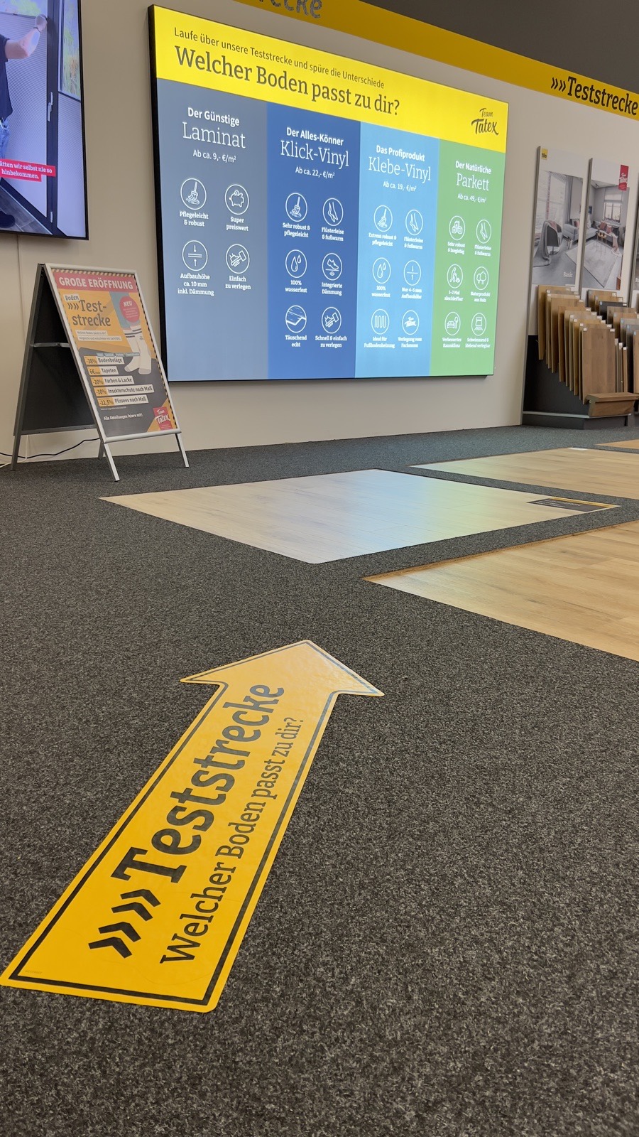 Die Tatex Teststrecke: gelber Pfeil auf altem Teppichboden führt zu Testflächen mit Laminat, Vinyl und Parkett